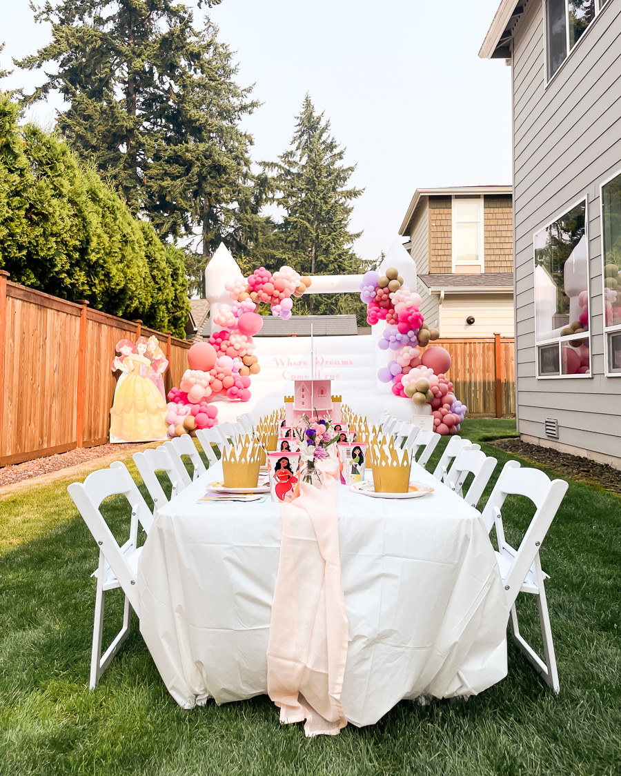 Disney princess birthday party decorations with white bounce house, pink and purple balloon garland, princess cardboard cutout, kids table setting with crown bowls, princess plates, and castle cupcake holder