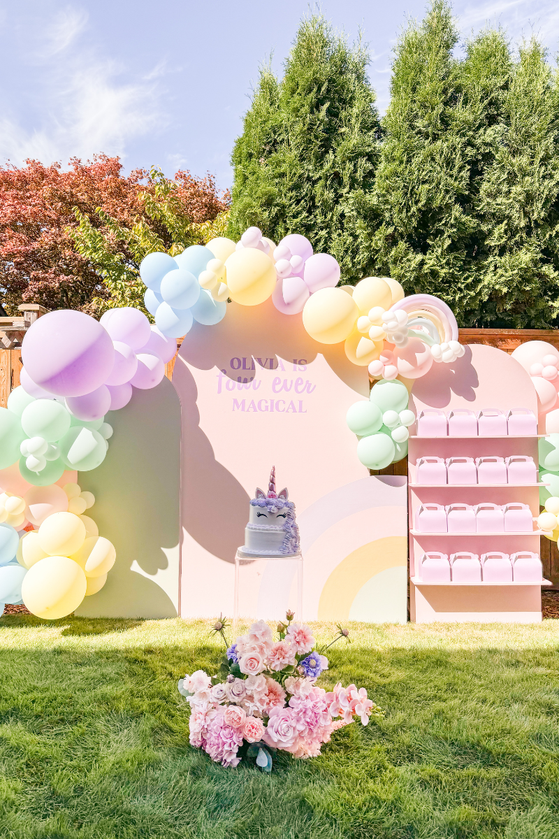 Arch backdrop for a kids unicorn birthday party with pastel balloon garland, lavender party favor boxes, and tiered unicorn cake on an acrylic cake stand